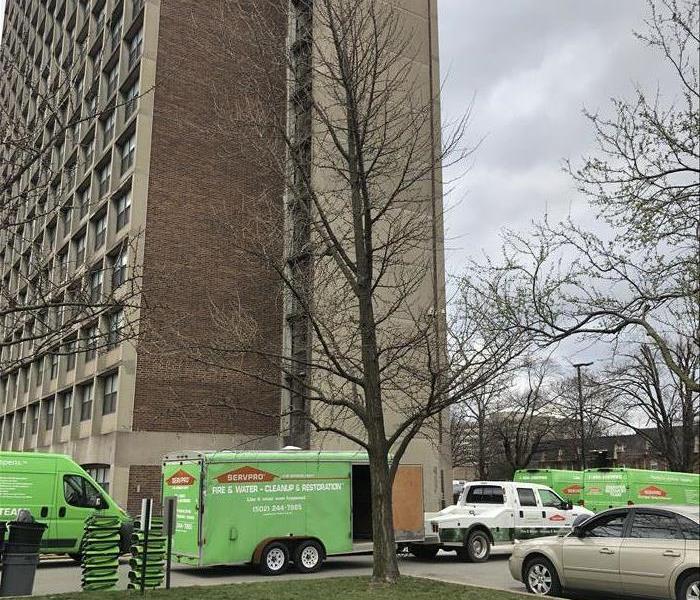 Multiple green SERVPRO vehicles parked in front of a tall apartment complex water damage